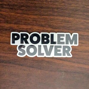Pode incluir: Um autocolante branco com as palavras "PROBLEM SOLVER" em negrito, com uma fonte sem serifa. A palavra "PROBLEM" é preta, e "SOLVER" está num gradiente de cinza. O autocolante está numa superfície castanha com grão de madeira.