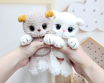 May include: Two crocheted cat toys, one white and one beige with brown ears. The beige cat has yellow ears and both cats have black eyes and pink noses.