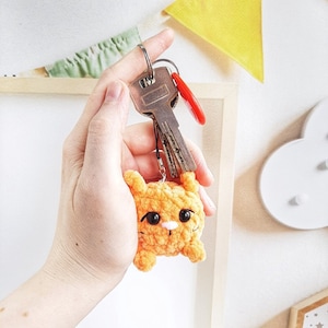 May include: A crocheted orange cat keychain with black eyes and a white nose attached to a silver key ring with two keys.