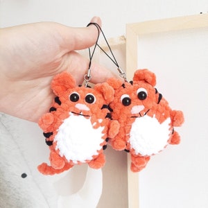 May include: Two orange and black crocheted tiger keychain charms with white faces and black eyes. The charms are attached to black cords with silver clasps.
