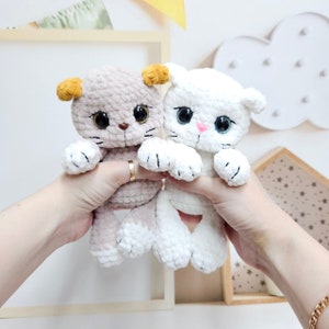 Peut inclure: Deux jouets en crochet en forme de chat, l'un blanc et l'autre beige avec des oreilles brunes. Le chat beige a des oreilles jaunes et les deux chats ont des yeux noirs et des nez roses.