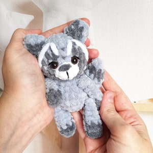 May include: A gray crocheted stuffed animal resembling a raccoon with white accents around the eyes and nose. The animal is being held in two hands.