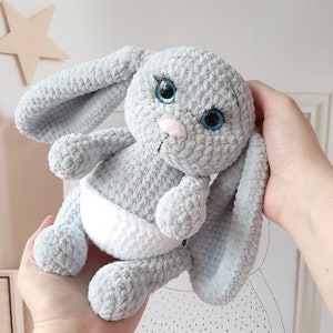 May include: A gray crocheted bunny toy with blue eyes and a white diaper. The bunny is sitting on a hand.