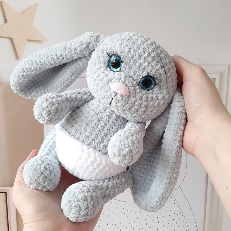 May include: A gray crocheted bunny toy with blue eyes and a white diaper. The bunny is sitting on a hand.