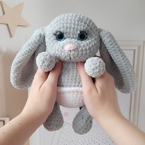 May include: A gray crocheted bunny with blue eyes and a white diaper. The bunny is being held by two hands.