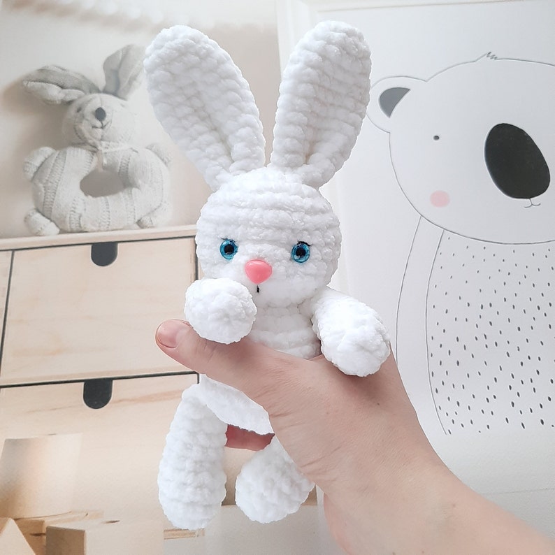 May include: A white crocheted bunny rabbit toy with blue eyes and a pink nose. The bunny is being held by a hand.
