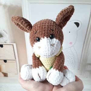 May include: A brown and white crocheted donkey plush toy with big brown eyes. The donkey is wearing a yellow crocheted vest.