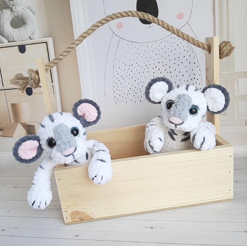 May include: Two white and gray crocheted tiger plushies are sitting in a wooden crate. The crate has a rope handle and is sitting on a white surface.