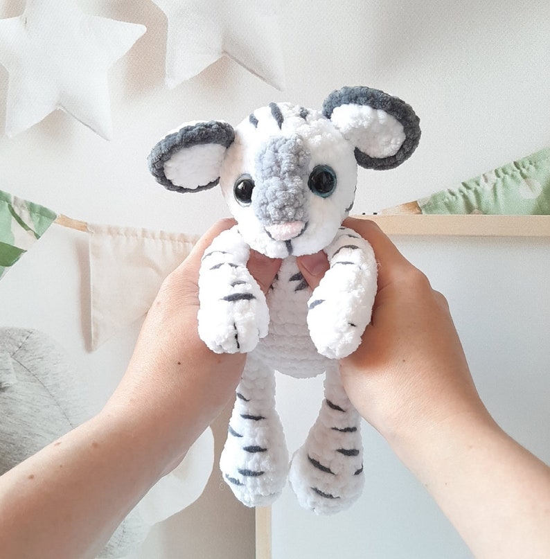May include: A white and gray crocheted tiger plush toy with black stripes. The toy has a gray nose and blue eyes.