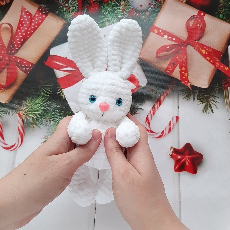 May include: A white crocheted bunny rabbit toy with blue eyes and a pink nose. The bunny is being held by a person's hands.