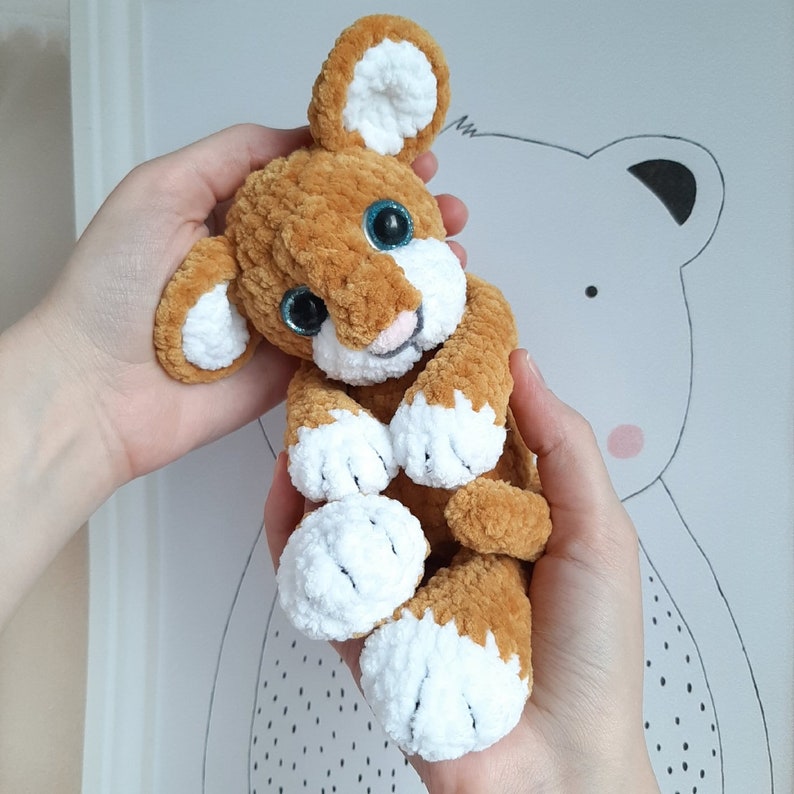 May include: A crocheted stuffed animal in the shape of a lion with brown fur and white accents. The lion has blue eyes and a pink nose.