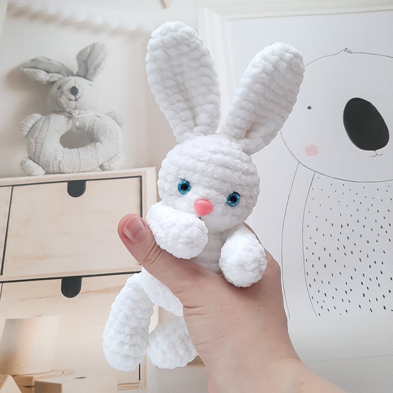 May include: A white crocheted bunny rabbit toy with blue eyes and a pink nose. The bunny is being held by a hand.