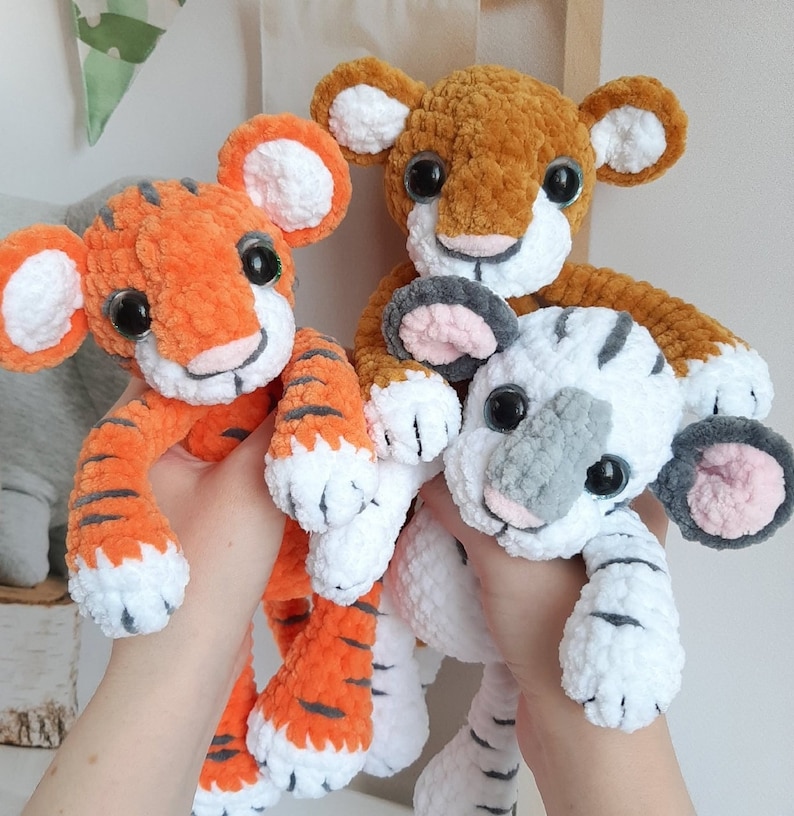 May include: Three crocheted stuffed animals: a tiger, a lion, and a white animal with gray accents. The tiger is orange and black with white stripes. The lion is brown with a white face and a white belly. The white animal has gray ears, a gray nose, and gray eyes.