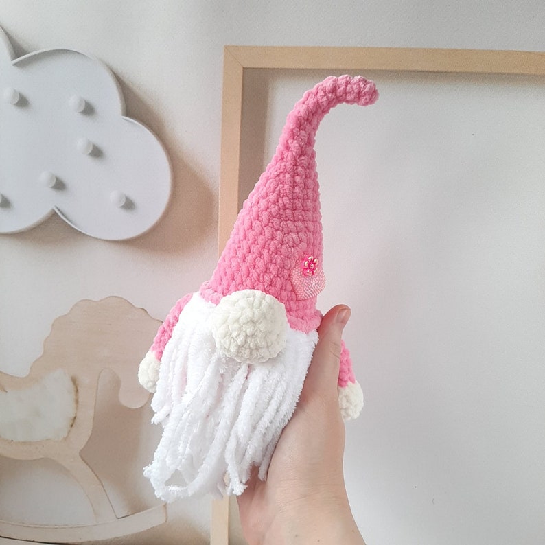 May include: A pink crocheted gnome with a white beard and pink glitter accents. The gnome is holding a pink flower.