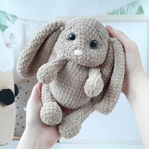 May include: A soft, crocheted plush bunny toy with long ears, brown fur, and large black eyes. The bunny has a pink nose and is held in a person's hands.