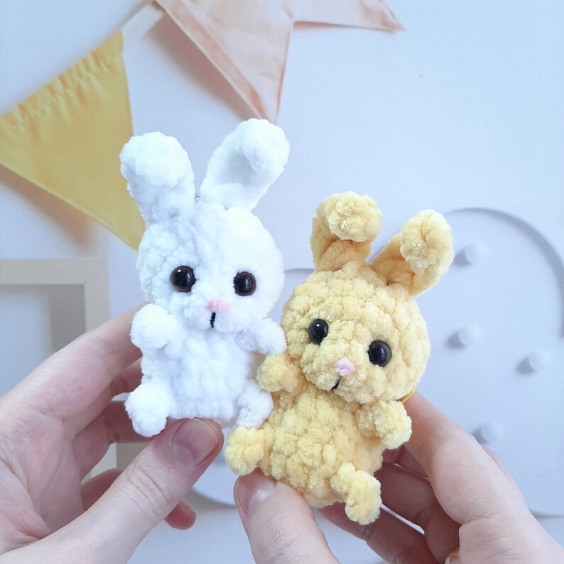 May include: Two crocheted bunny plushies, one white and one yellow. The white bunny has black eyes and a pink nose. The yellow bunny has black eyes and a pink nose.