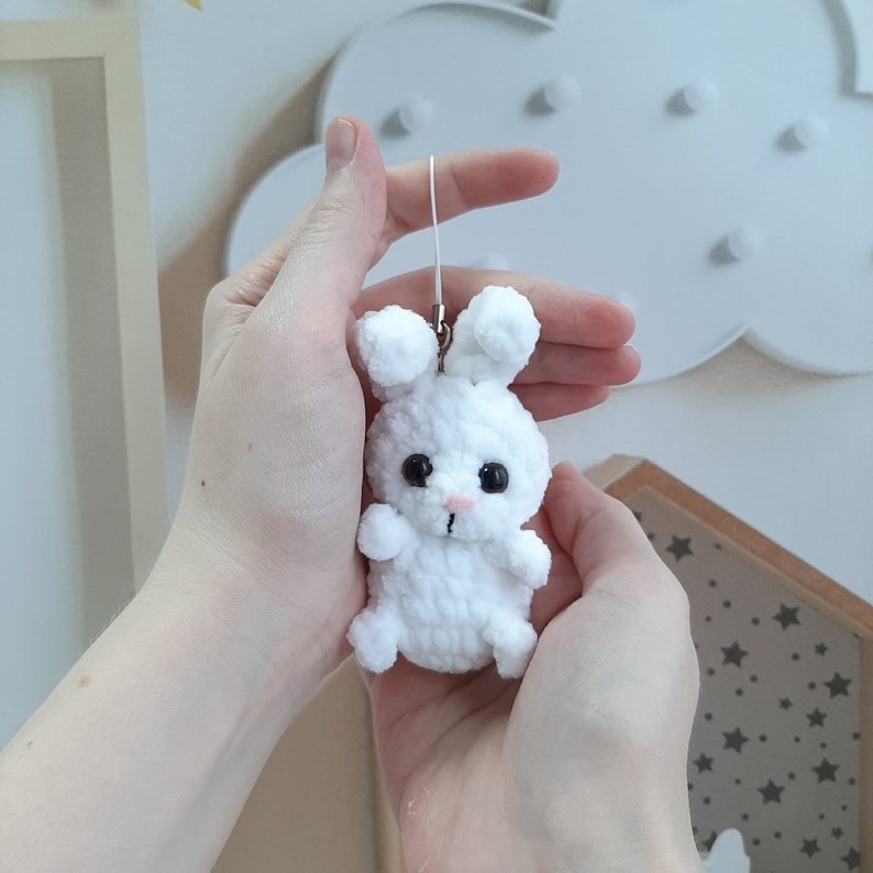 May include: A white crocheted bunny keychain with black eyes and a pink nose. The bunny is being held in a person's hands.