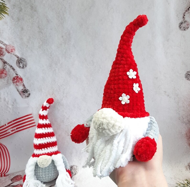 May include: Two crocheted gnomes, one with a red and white striped hat and the other with a red hat with white flowers. Both gnomes have white yarn beards and gray bodies.