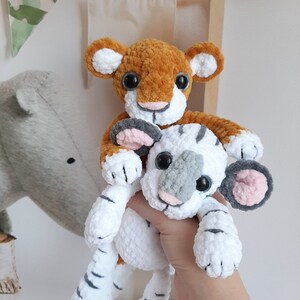 May include: A crocheted tiger plush toy with orange and brown fur, riding on a white crocheted tiger plush toy with gray and white stripes. Both toys have black eyes and a pink nose.