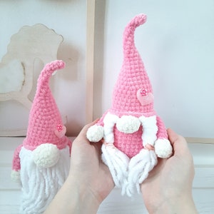 May include: Two pink crocheted gnomes with white beards and pink hearts. The larger gnome is holding a pink heart in its hand.