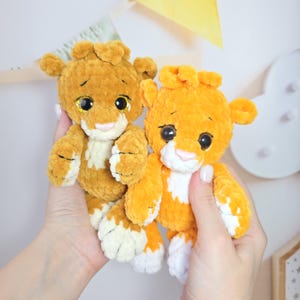 May include: Two crocheted lion plushies, one brown and one orange, with white accents on their paws and faces. They have large, sparkly eyes and a sweet expression.