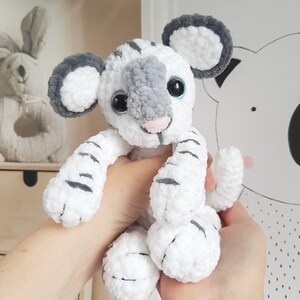 May include: White and gray crocheted tiger plush toy with blue eyes. The toy is sitting in a person's hands.