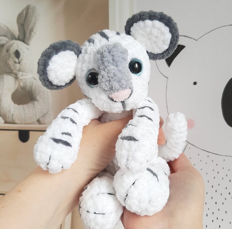 May include: White and gray crocheted tiger plush toy with blue eyes. The toy is sitting in a person's hands.