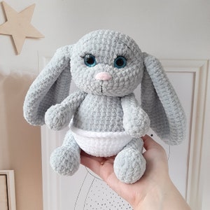 May include: A gray crocheted bunny with blue eyes and a white diaper. The bunny is sitting on a hand.