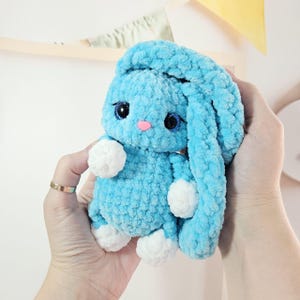 May include: A light blue plush bunny with long ears, white paws, and a pink nose. The handmade stuffed animal has large, expressive eyes and is held in two hands. The bunny is a soft, cuddly toy.