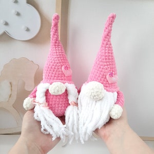May include: Two pink crocheted gnomes with white beards and pink hearts on their hats. The gnomes are holding hands and are sitting on a white surface.