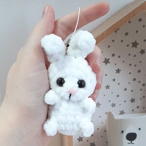 May include: White crocheted bunny keychain with black eyes and a pink nose. The bunny is holding a white string.