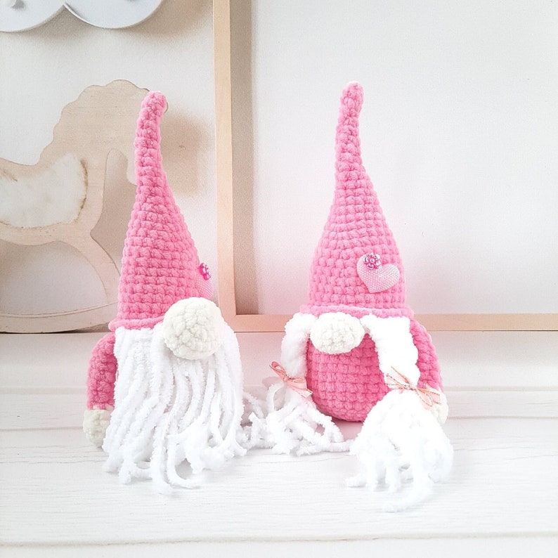 May include: Two pink crocheted gnomes with white yarn beards and pink hearts on their hats. The gnomes are sitting on a white surface.