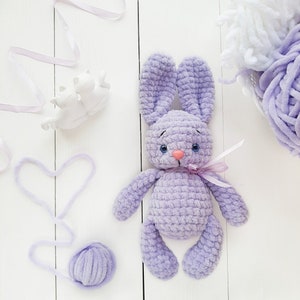 May include: A lavender crocheted bunny toy with a pink nose and blue eyes. The bunny is sitting on a white surface with a ball of lavender yarn and a white ribbon.