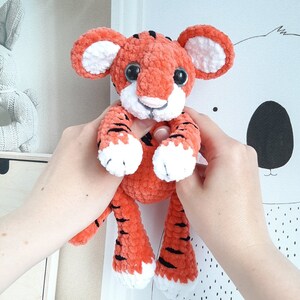 May include: A crocheted tiger plush toy with orange and black stripes, white paws, and black eyes. The toy is being held by a person's hands.