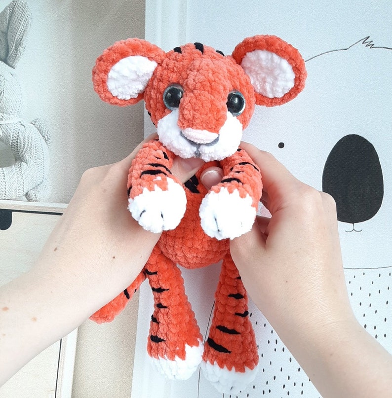 May include: A crocheted tiger plush toy with orange and black stripes, white paws, and black eyes. The toy is being held by a person's hands.