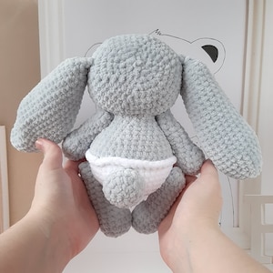 May include: A gray crocheted bunny with long floppy ears and a white crocheted diaper. The bunny is sitting on a white surface.