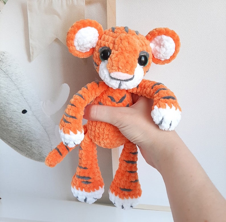 May include: A crocheted orange and white tiger plush toy with black stripes. The toy has a fluffy texture and large, round eyes.