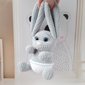 May include: A gray crocheted bunny rabbit toy with blue eyes and a white diaper. The bunny is hanging from a hand.