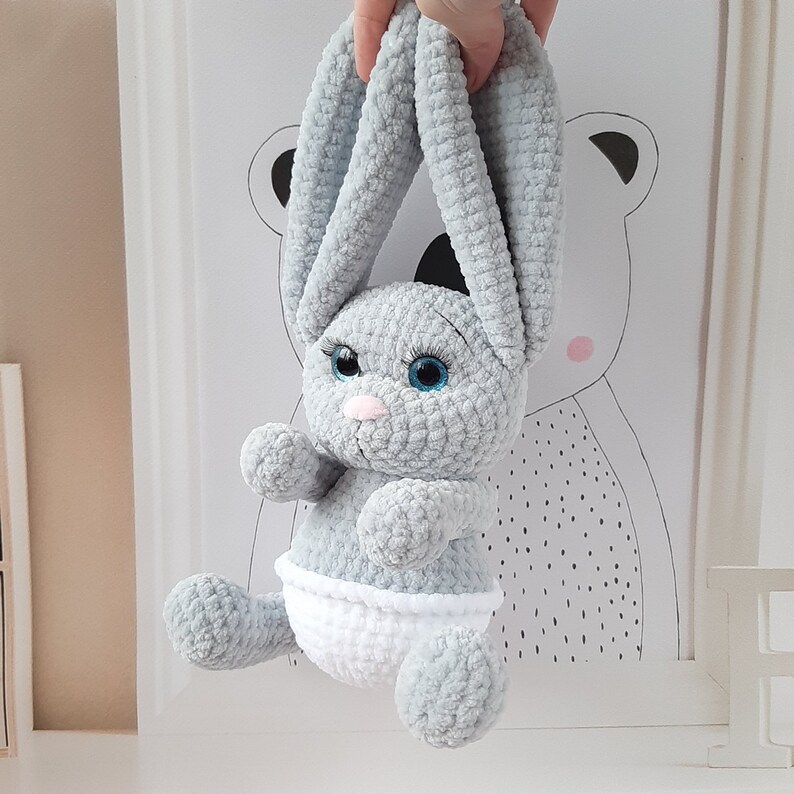 May include: A gray crocheted bunny rabbit toy with blue eyes and a white diaper. The bunny is hanging from a hand.