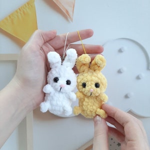 May include: Two crocheted bunny keychains, one white and one yellow, with black eyes and pink noses. The keychains are hanging from a white string.