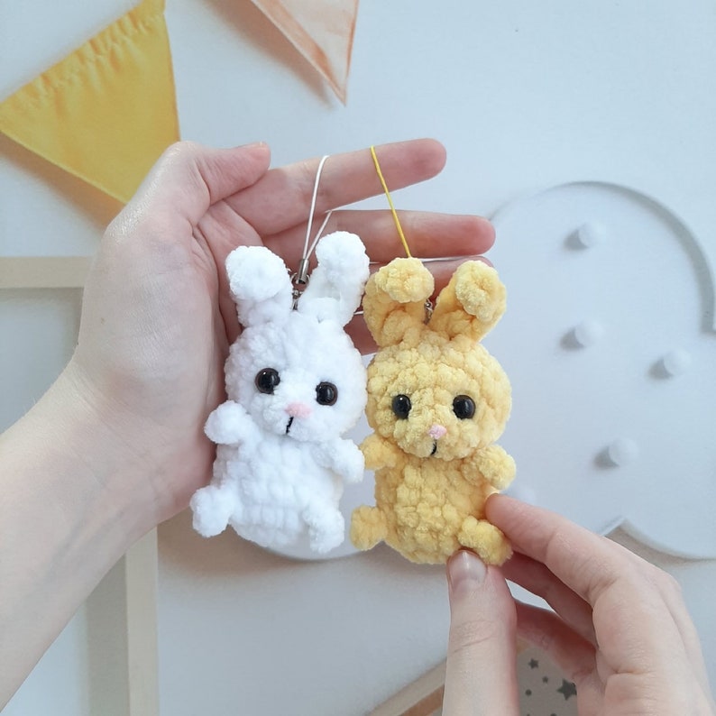 May include: Two crocheted bunny keychains, one white and one yellow, with black eyes and pink noses. The keychains are hanging from a white string.