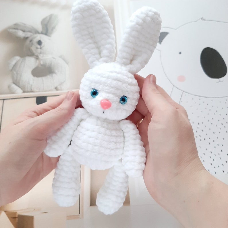 May include: A white crocheted bunny rabbit toy with blue eyes and a pink nose. The bunny is being held by two hands.