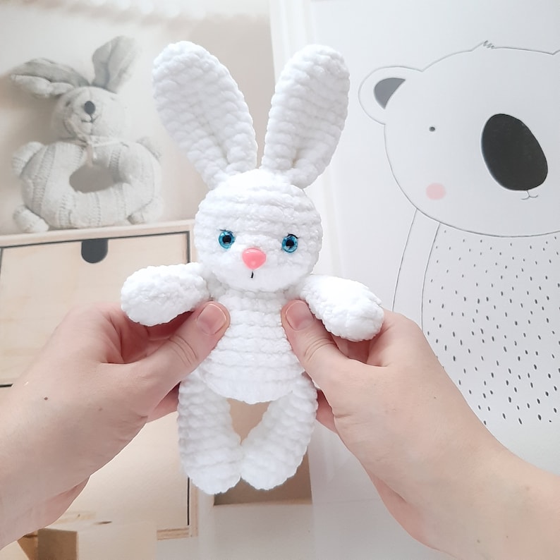 May include: A white crocheted bunny rabbit toy with blue eyes and a pink nose. The bunny is being held by a person's hands.