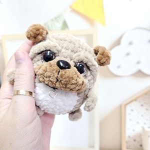 Puede incluir: Un perro pug de peluche hecho a mano con cuerpo beige, orejas y hocico marrones, y ojos grandes y brillantes. El pug tiene una nariz negra y un pequeño dije en forma de corazón en su collar. El juguete es suave y tierno.