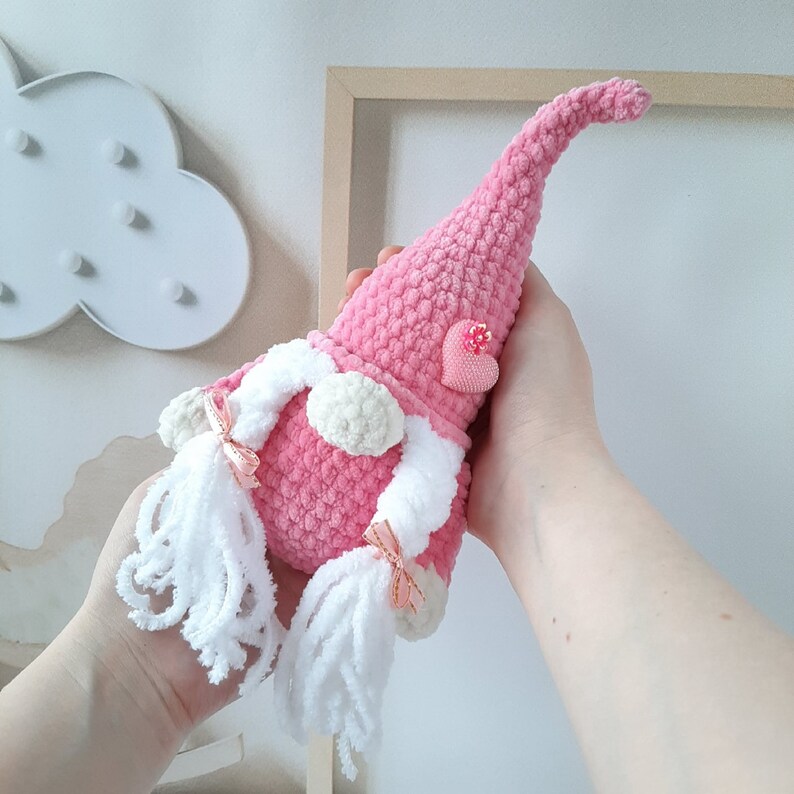 May include: A pink crocheted gnome with a white beard and pink heart embellishment. The gnome has a long pink hat and white yarn braids.