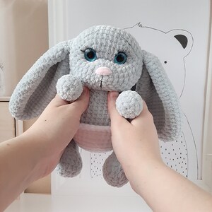 May include: A gray crocheted bunny rabbit toy with blue eyes and a pink belly. The bunny is being held by two hands.