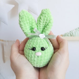 May include: A green crocheted bunny with a white flower and silver ribbon. The bunny has black eyes and a pink nose.