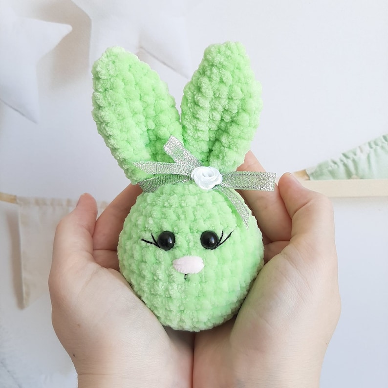 May include: A green crocheted bunny with a white flower and silver ribbon. The bunny has black eyes and a pink nose.