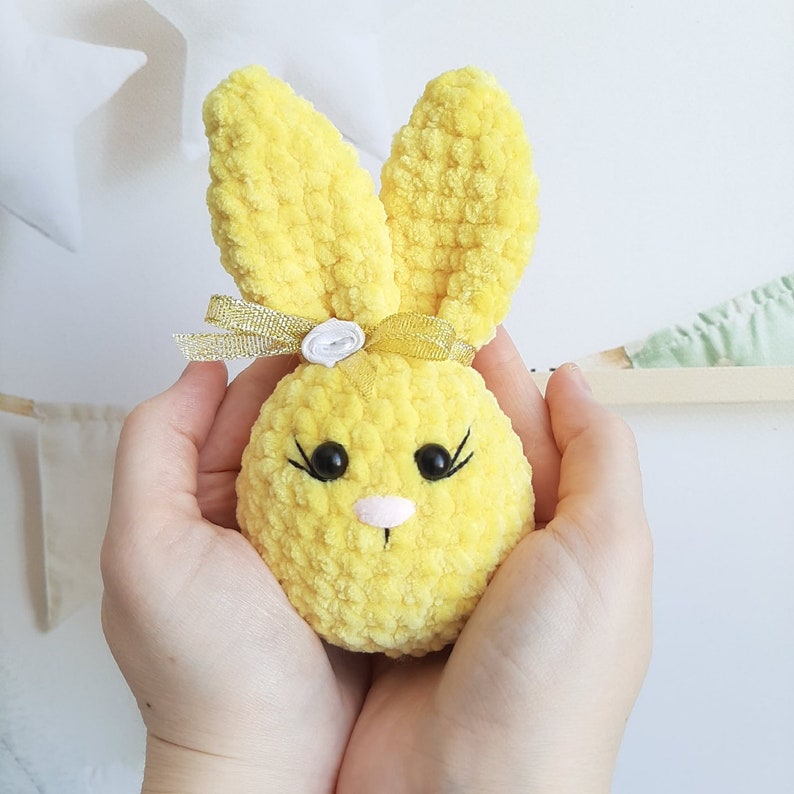 May include: A yellow crocheted bunny with a white flower and a gold ribbon. The bunny has black eyes and a pink nose.