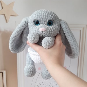 May include: A gray crocheted bunny with blue eyes and a white diaper. The bunny is being held by a hand.